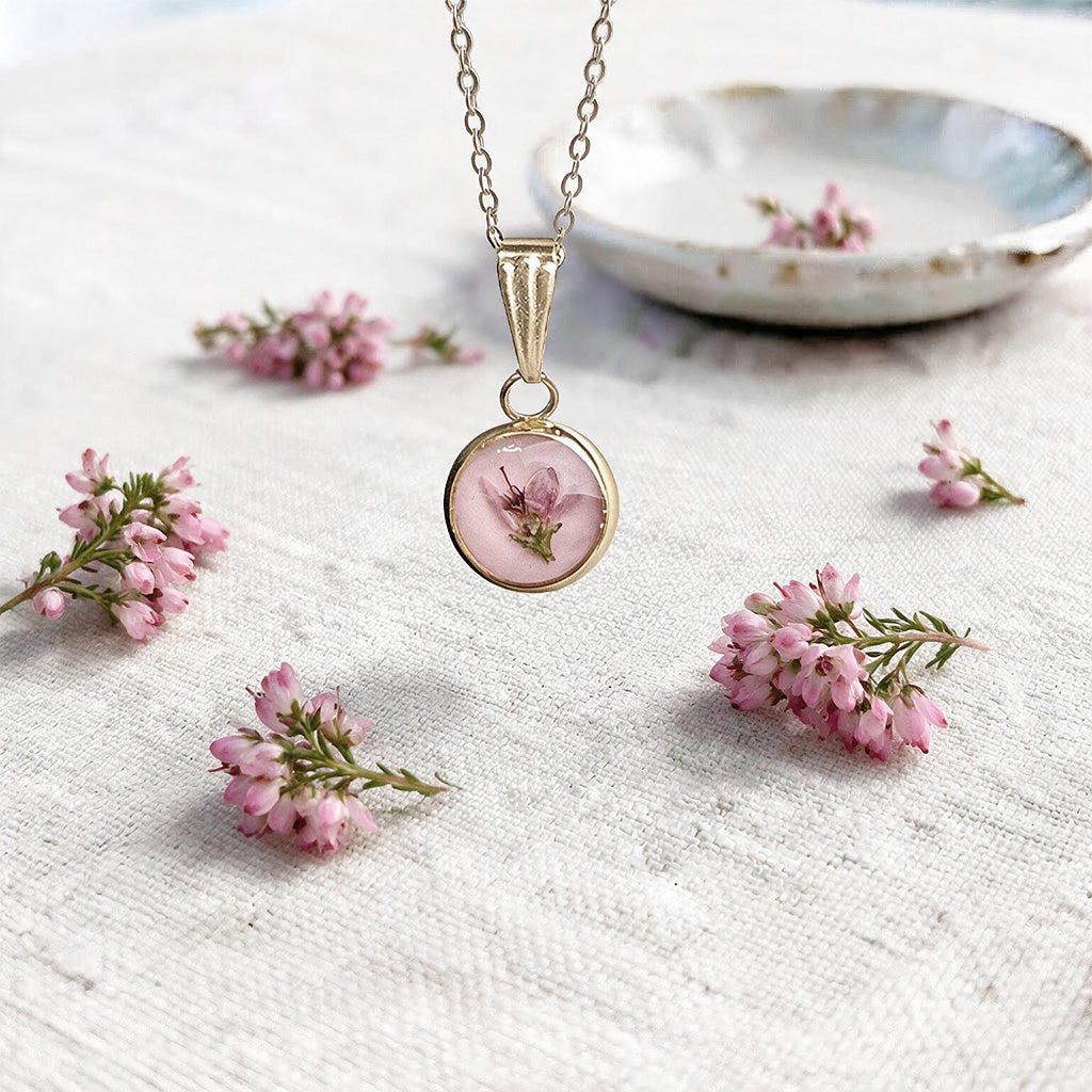 Heather flower necklace with a real pressed heather blossom preserved in clear resin, photographed on natural linen surrounded by fresh pink heather sprigs. Handcrafted in Finland.
