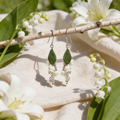 Lily of the Valley Earrings displayed on a birch branch among lily of the valley flowers and white lilies in bright natural daylight. Handmade in Finland by byMinta.