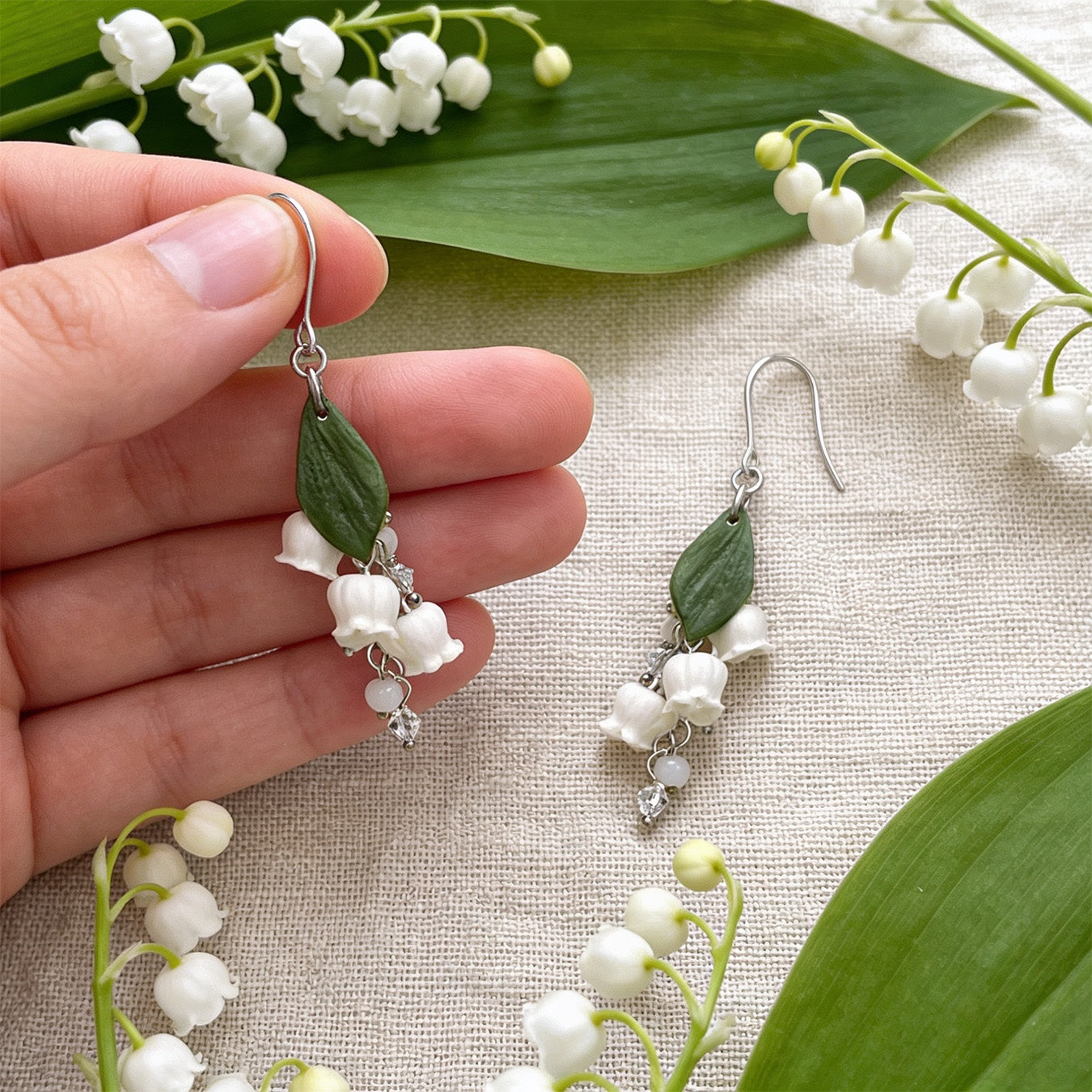 Lily of the Valley Earrings photographed with fresh lily of the valley flowers and green leaves in soft natural light, with one earring gently held by hand for scale. Handmade in Finland by byMinta.