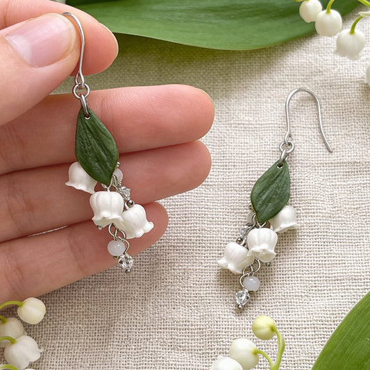 Lily of the Valley Earrings photographed with fresh lily of the valley flowers and green leaves in soft natural light, with one earring gently held by hand for scale. Handmade in Finland by byMinta.