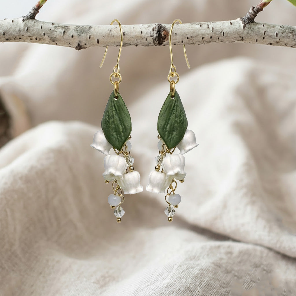 Handcrafted Lily of the Valley flower earrings displayed on a birch branch over natural linen fabric.