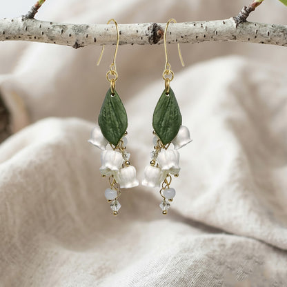Handcrafted Lily of the Valley flower earrings displayed on a birch branch over natural linen fabric.