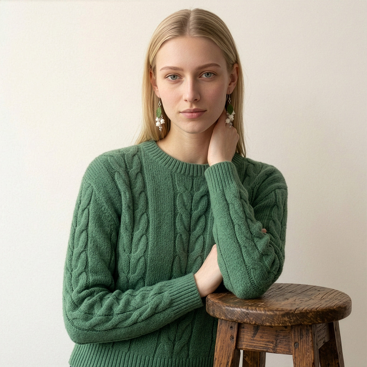 Blonde woman wearing delicate white floral earrings by byMinta, paired with a soft forest green cable-knit sweater. She poses indoors against a neutral background, resting one hand lightly at her neck beside a rustic wooden stool. The calm green tones and natural styling create a timeless Nordic look inspired by the quiet beauty of the forest.