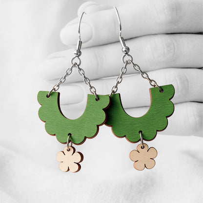 Green and birch Kiikku (Swing) plywood earrings handmade in Finland, photographed hanging against a black-and-white hand background.
