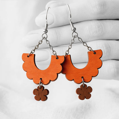 Orange and Walnut Kiikku (Swing) plywood earrings handmade in Finland, photographed hanging against a black-and-white hand background.