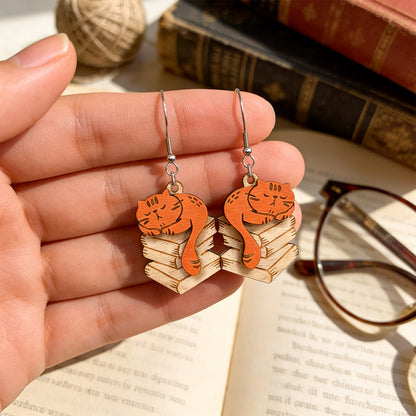 Hand holding handmade Library Cat earrings for size reference over an open book, with vintage books, reading glasses and a ball of yarn in the background. Lightweight Finnish birch plywood cat earrings photographed in warm natural light with a cozy book lover atmosphere.