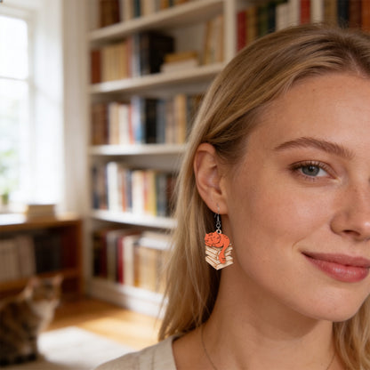 Close-up portrait of a Scandinavian woman wearing handmade Library Cat earrings in a cozy home library. Lightweight Finnish birch plywood cat earrings photographed in warm natural light with bookshelves and a softly blurred cat in the background.