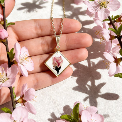 Hand holding a handmade resin necklace with a real cherry blossom for size reference, surrounded by delicate pink cherry blossoms in bright natural daylight. Botanical jewelry photographed with a soft and airy spring atmosphere.
