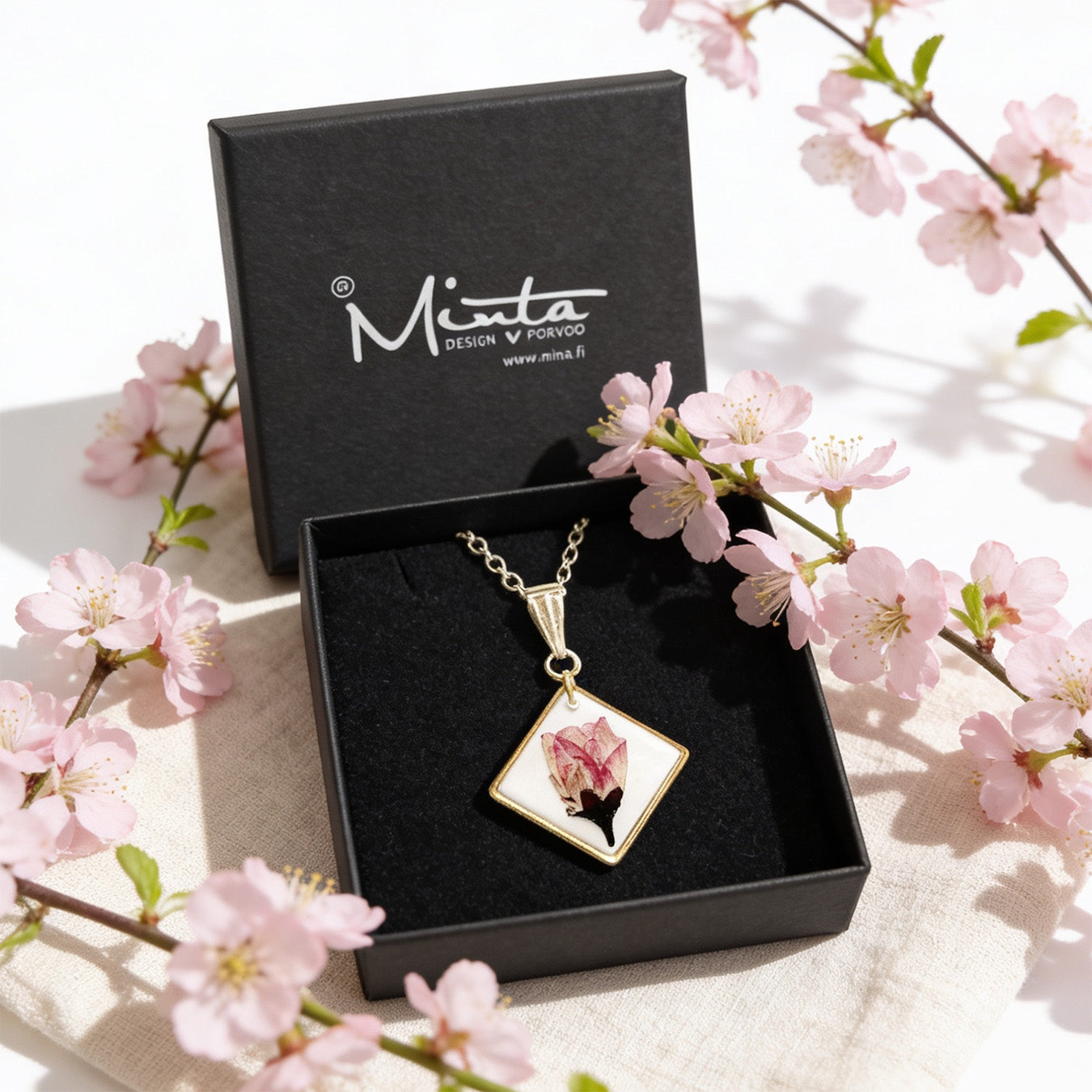 Handmade resin necklace with a real cherry blossom displayed in a black jewelry box surrounded by delicate pink cherry blossoms. Botanical jewelry photographed in bright natural daylight with a soft and airy spring atmosphere.