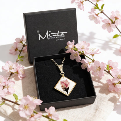 Handmade resin necklace with a real cherry blossom displayed in a black jewelry box surrounded by delicate pink cherry blossoms. Botanical jewelry photographed in bright natural daylight with a soft and airy spring atmosphere.