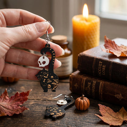 Hand holding black cat earrings with skull charms, engraved Halloween earrings handmade in Finland.