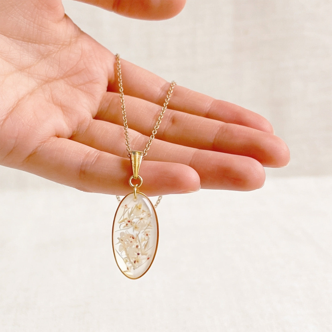 Hand holding a handmade botanical necklace for size reference, featuring real pressed cow parsley flowers preserved in clear resin. Oval gold-tone pendant on a delicate chain against a soft linen background in bright natural daylight.