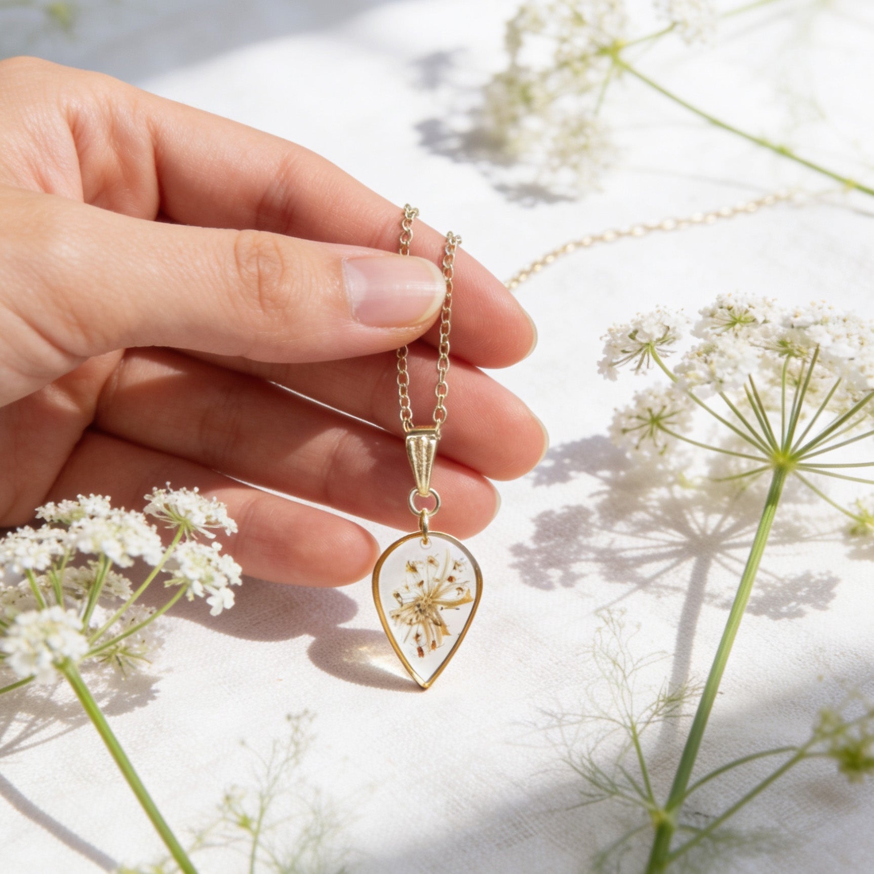 Hand holding a handmade botanical necklace for size reference, featuring real pressed cow parsley flowers preserved in clear resin. Surrounded by delicate white cow parsley blooms in bright natural daylight with a light and airy summer atmosphere.