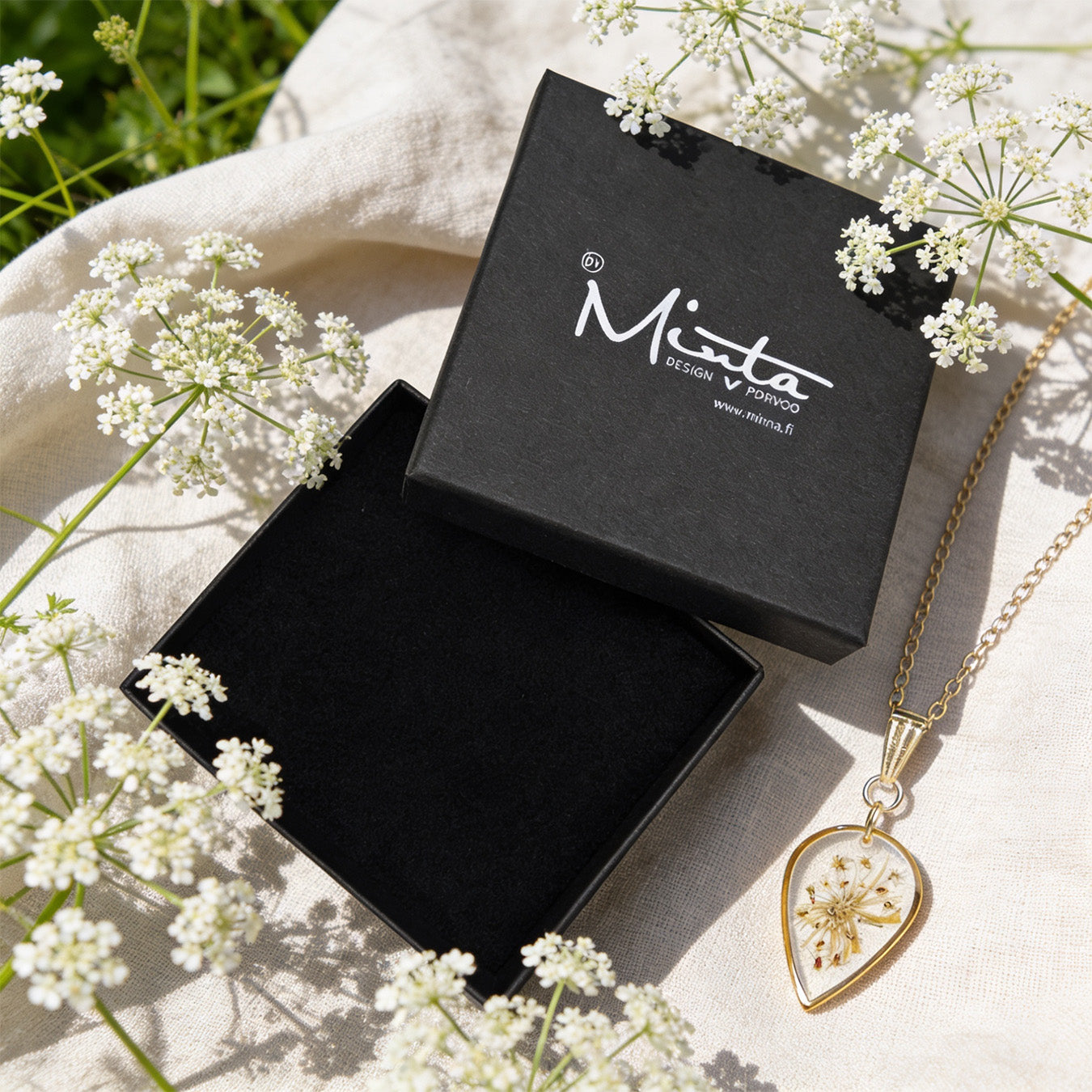 Handmade botanical necklace with real pressed cow parsley flowers preserved in clear resin, displayed beside a black byMinta jewelry box. Surrounded by delicate white cow parsley blooms and soft linen in bright natural daylight with a fresh Nordic summer atmosphere.