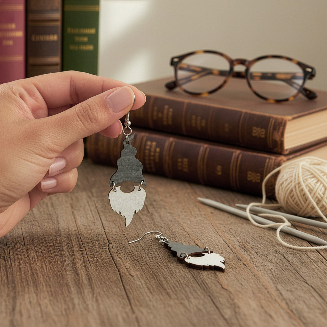 Hand holding a silver Christmas gnome earring on a wooden table with vintage books, glasses, knitting needles, and soft white yarn. Handmade in Finland by byMinta.