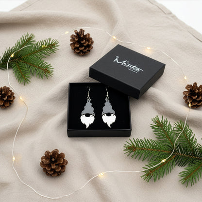 Gray and white Christmas gnome earrings presented in a gift box with spruce branches, pinecones, and warm fairy lights in a festive holiday setting. Handmade in Finland by byMinta.
