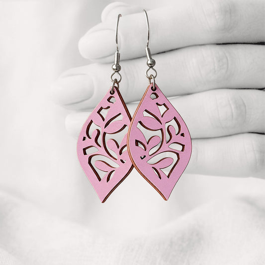 Mauve Köynnös plywood earrings with intricate floral cut-out details, made in Finland, photographed hanging from a hand in black and white against a softly blurred light background.