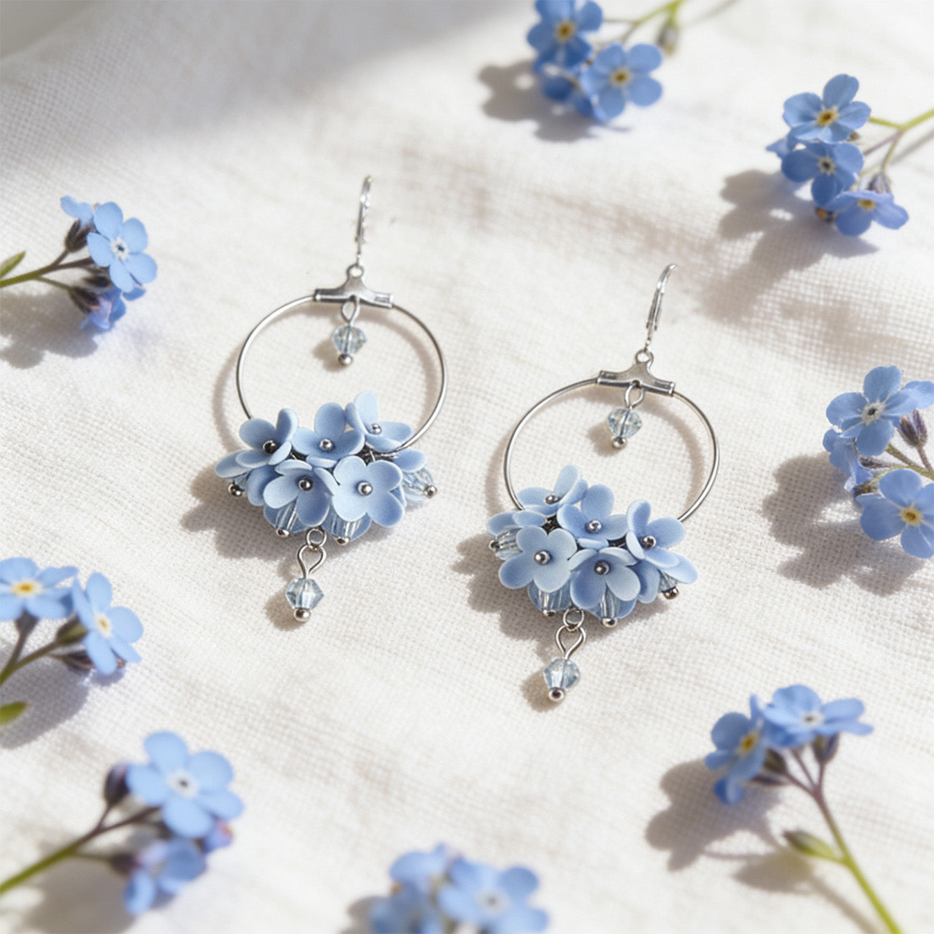 Light Blue Flower Vine Hoop Earrings resting on linen among blooming forget-me-nots in bright natural daylight. Handmade in Finland by byMinta.