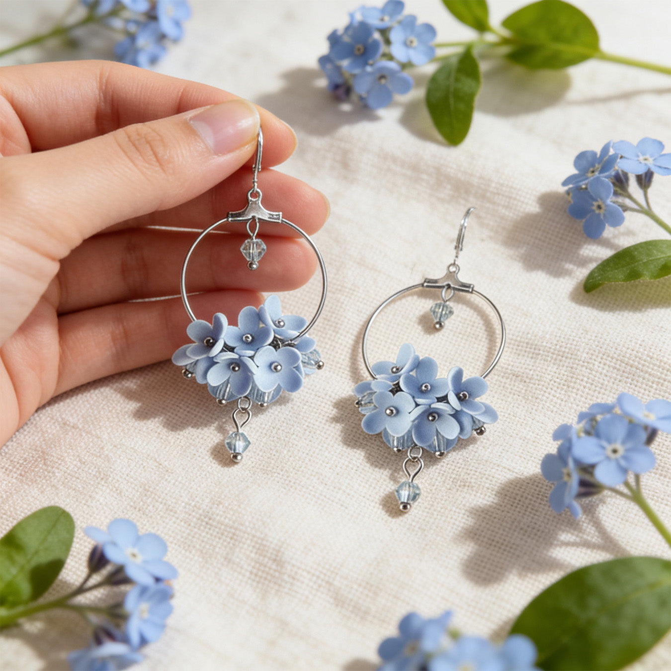 Light Blue Flower Vine Hoop Earrings photographed with forget-me-nots in soft natural light, with one earring gently held by hand for scale. Handmade in Finland by byMinta.