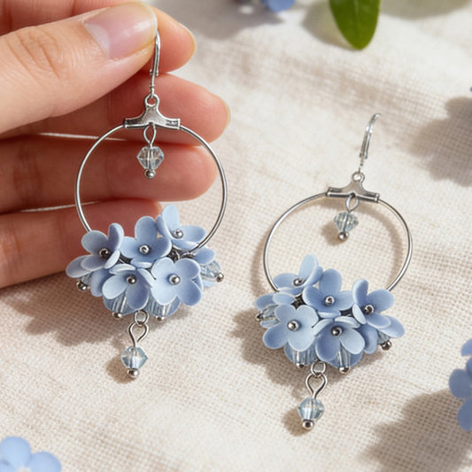 Light Blue Flower Vine Hoop Earrings photographed with forget-me-nots in soft natural light, with one earring gently held by hand for scale. Handmade in Finland by byMinta.