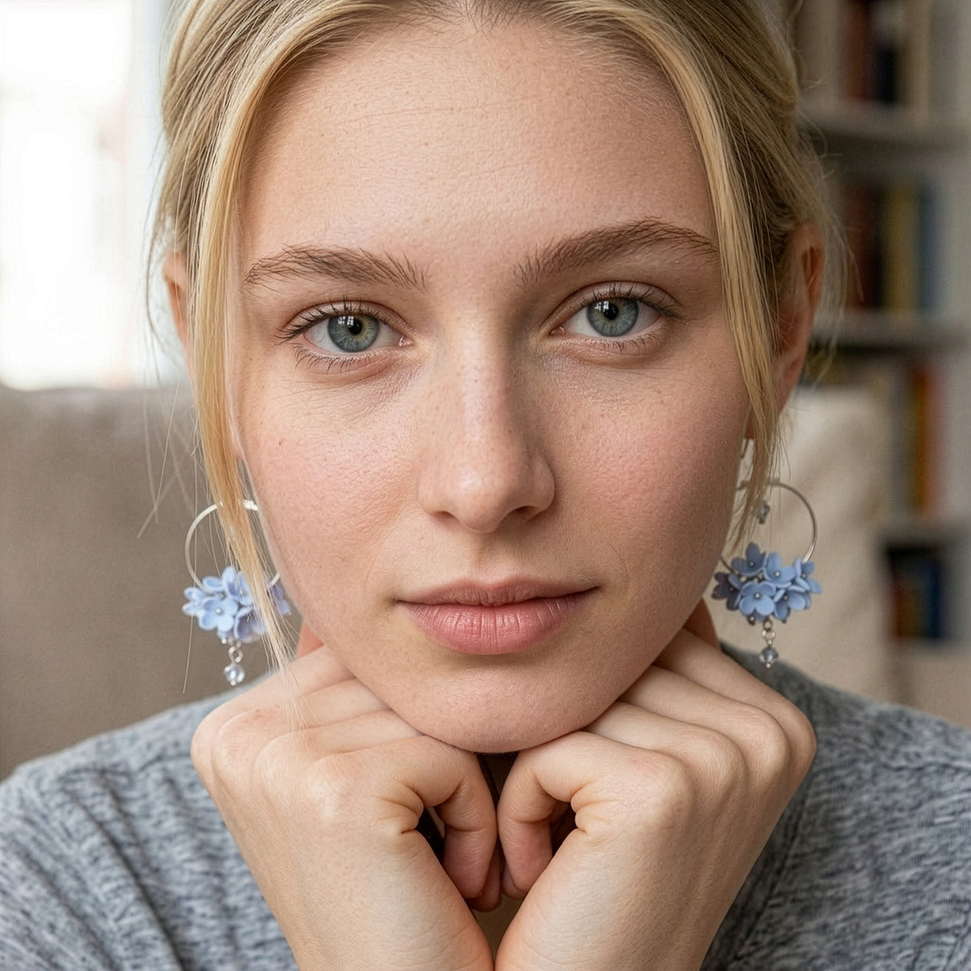 Close-up portrait of a blonde woman wearing light blue floral hoop earrings by byMinta. She rests her chin on her hands and looks directly into the camera with a calm, natural expression. The soft indoor lighting and neutral background keep the focus on the delicate handmade earrings, inspired by the gentle colors of the summer sky and northern flowers.