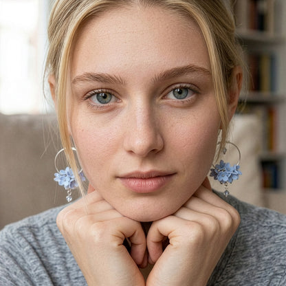 Close-up portrait of a blonde woman wearing light blue floral hoop earrings by byMinta. She rests her chin on her hands and looks directly into the camera with a calm, natural expression. The soft indoor lighting and neutral background keep the focus on the delicate handmade earrings, inspired by the gentle colors of the summer sky and northern flowers.