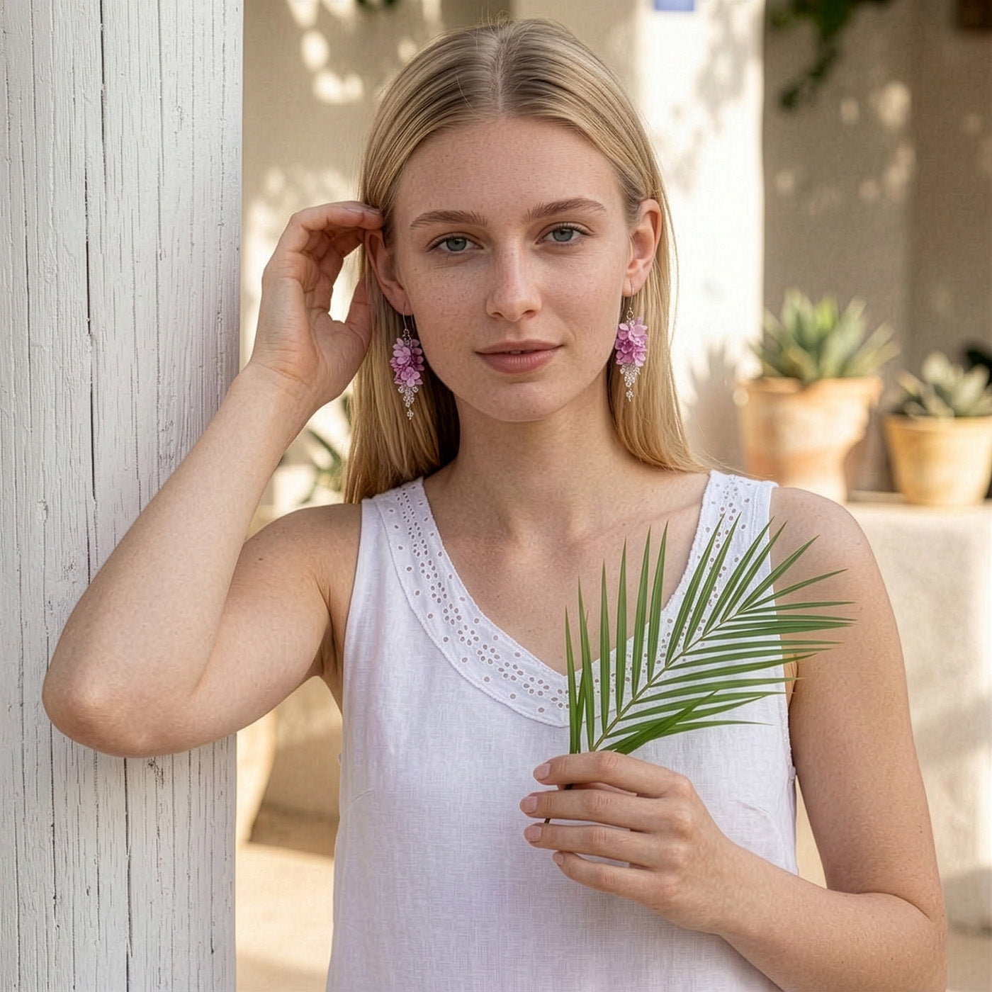 Blonde woman wearing delicate pink floral earrings by byMinta, standing in soft natural light beside a white column. She is dressed in a simple white sleeveless top and holds a green palm leaf in her hand. The warm, airy setting and relaxed pose create a fresh and elegant summer look inspired by blooming flowers and handcrafted details.