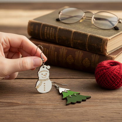 Hand holding the snowman earring on a wooden table with vintage books and red yarn, with the Christmas tree earring beside it. Handmade in Finland by byMinta.