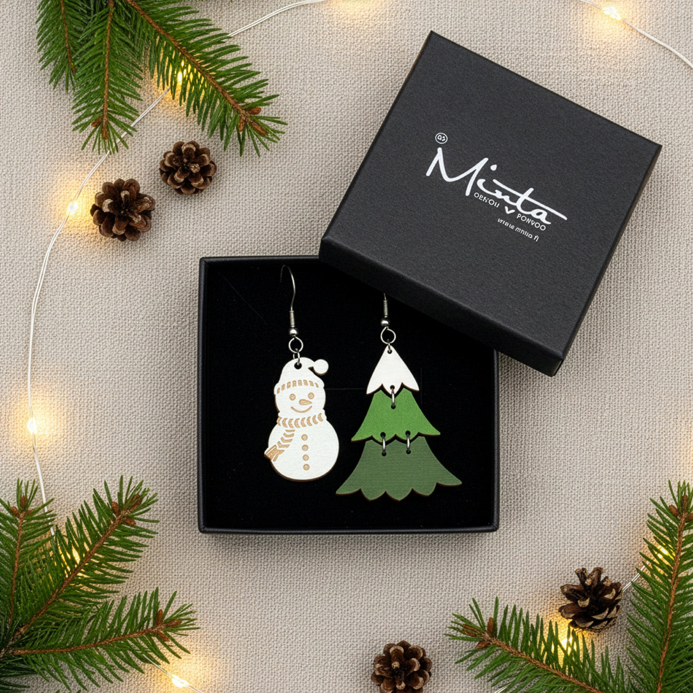 Snowman and Christmas tree earrings presented in a gift box with pine branches, pinecones, and warm fairy lights, creating a festive holiday atmosphere. Handmade in Finland by byMinta.
