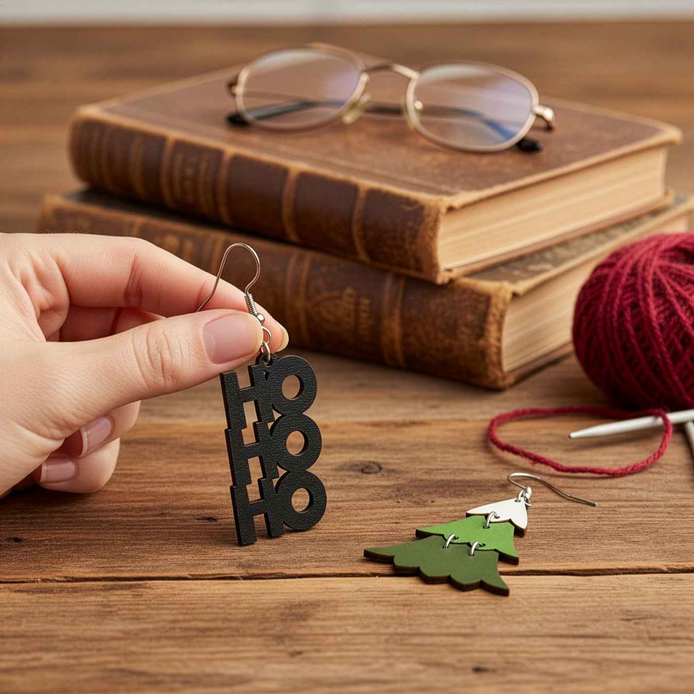 Hand holding the HO HO HO earring on a wooden table with vintage books, glasses, knitting needles, and burgundy yarn, with the Christmas tree earring beside it. Handmade in Finland by byMinta.