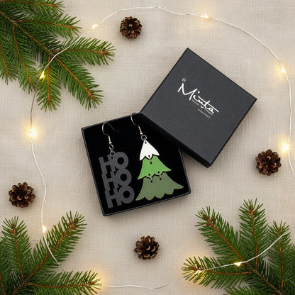 HO HO HO and Christmas tree earrings presented in a gift box with pine branches, pinecones, and warm fairy lights, creating a festive holiday atmosphere. Handmade in Finland by byMinta.
