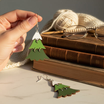 Hand holding a Christmas tree earring beside vintage books, glasses, yarn, and a knitted scarf in a warm winter setting. Handmade in Finland by byMinta.