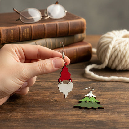 Hand holding a red gnome earring on a wooden table with vintage books, glasses, and soft white yarn, with the Christmas tree earring beside it. Handmade in Finland by byMinta.