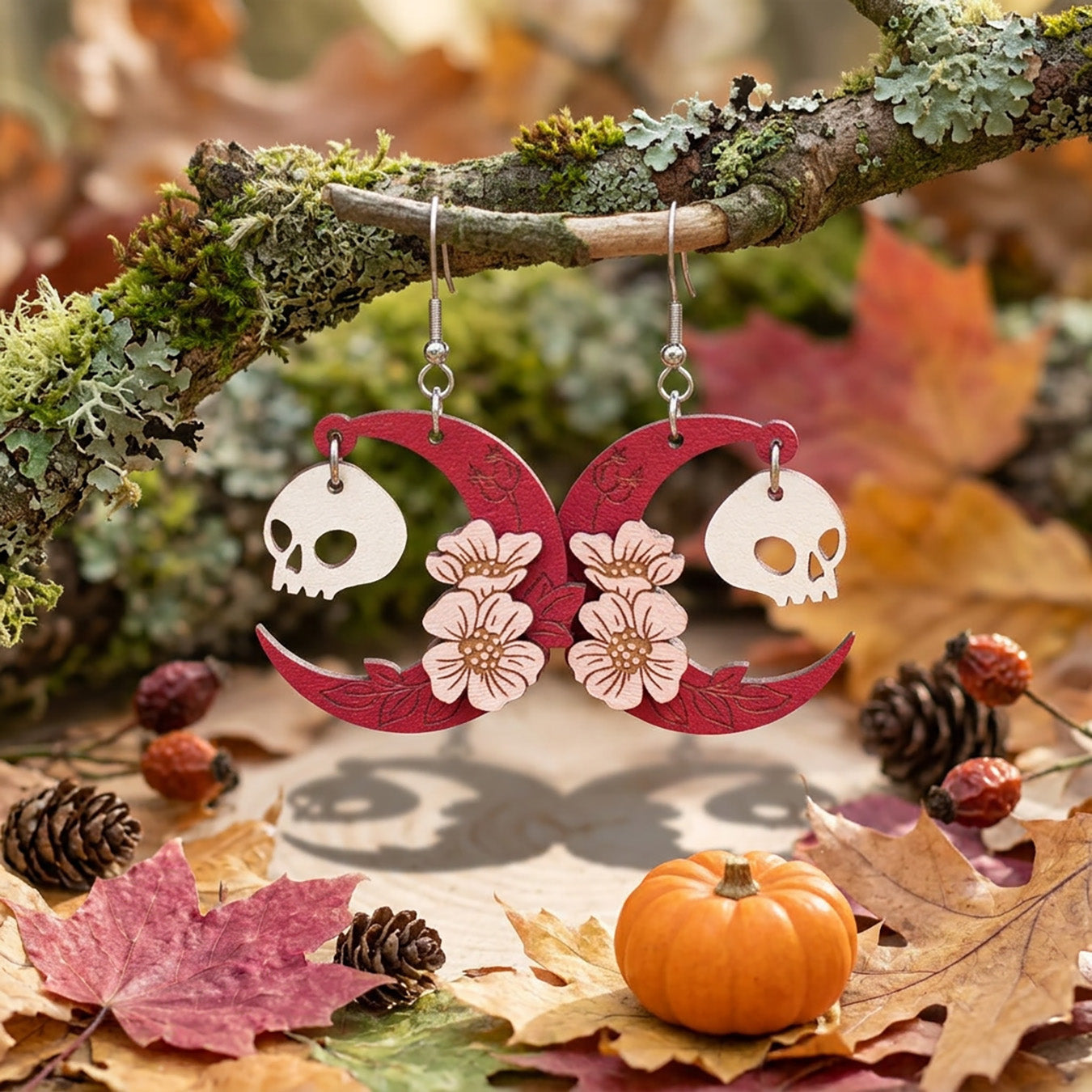 Pink crescent moon earrings with floral details and skull charms hanging from branch in autumn forest