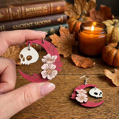 Hand holding pink crescent moon Halloween earrings with skull charms and flowers on autumn table