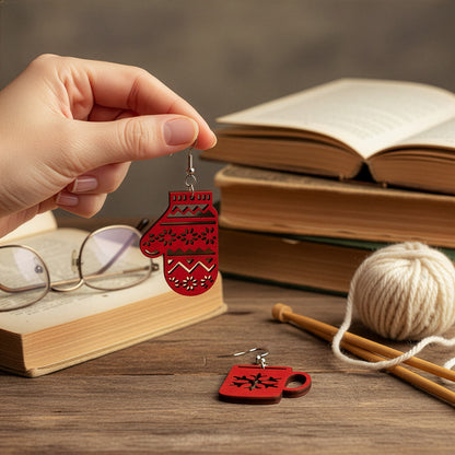 Hand holding the red mitten earring on a wooden table with books, knitting yarn, and reading glasses, with the mug earring beside it. Handmade in Finland by byMinta.