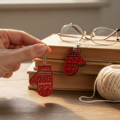 Hand holding a red mitten earring on a wooden table with books, glasses, and yarn, creating a cozy winter atmosphere. Handmade in Finland by byMinta.