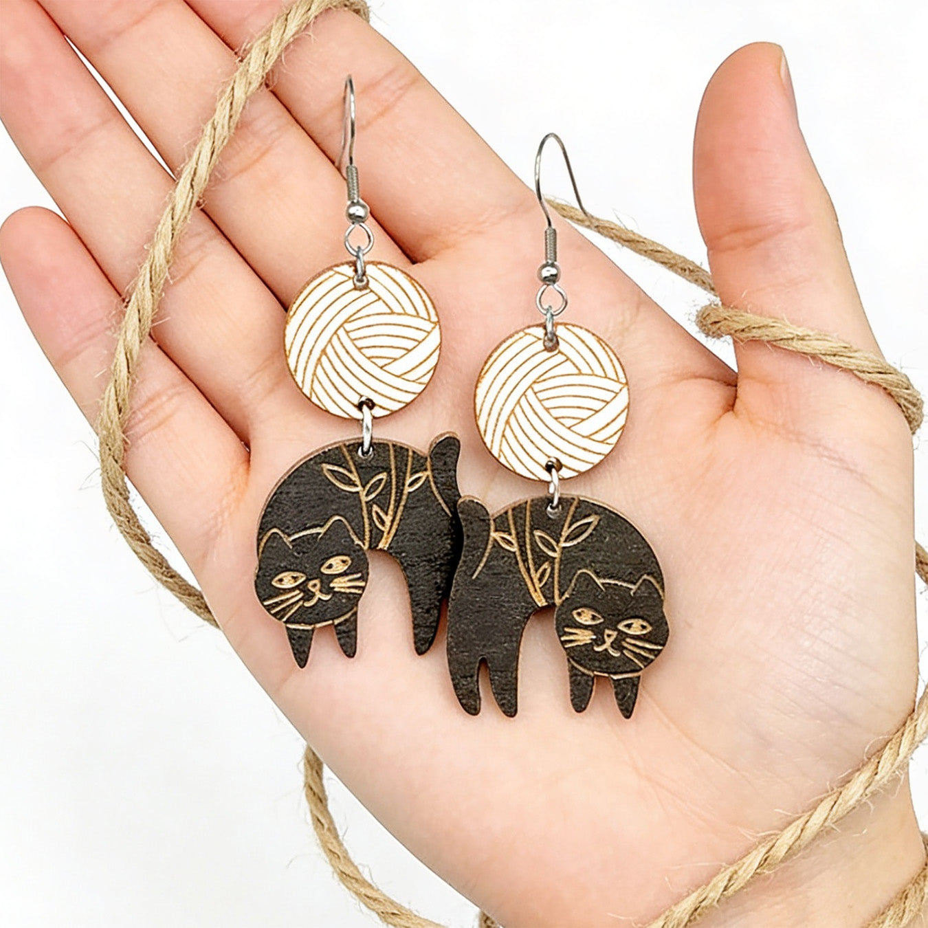 Hand holding handmade black cat earrings for size reference, wrapped in natural rope. Lightweight Finnish birch plywood earrings with engraved cat and botanical details in soft natural light.