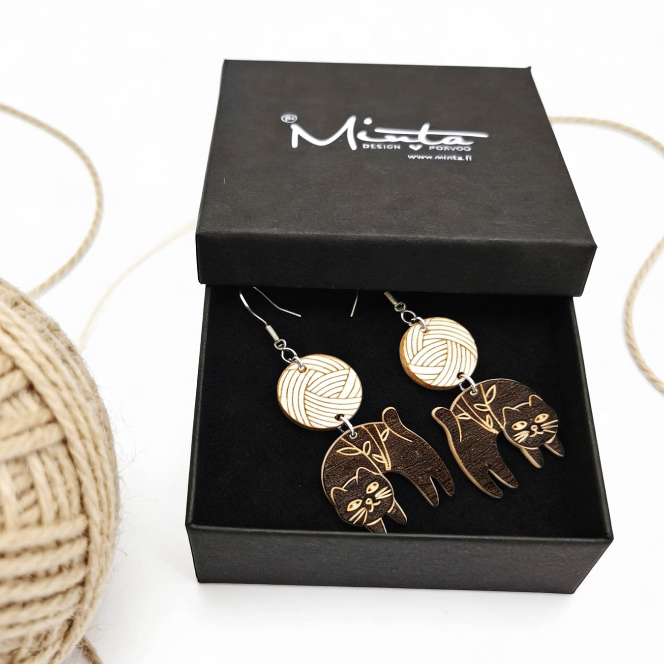 Handmade black cat earrings displayed in a black jewelry gift box beside a ball of natural yarn. Lightweight Finnish birch plywood earrings with engraved cat details, styled in a cozy Nordic craft atmosphere.
