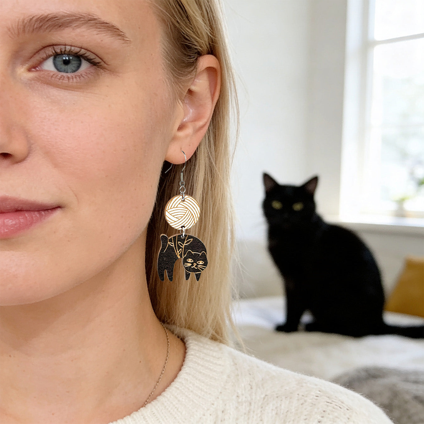 Close-up portrait of a Scandinavian woman wearing handmade black cat earrings with geometric wooden details. A black cat sits softly blurred in the background, creating a cozy Nordic home atmosphere with soft natural daylight.