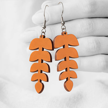 Orange Lehdykkä (Leaflet) plywood earrings made in Finland, photographed hanging from a hand in black and white against a softly blurred light background.