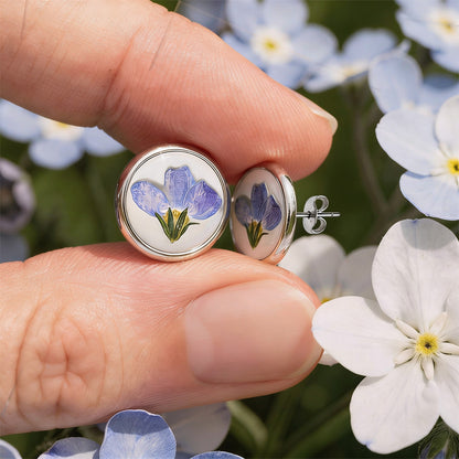 Forget-me-not stud earrings with real blue flowers preserved in resin, photographed between fingers among soft woodland flowers in natural daylight.