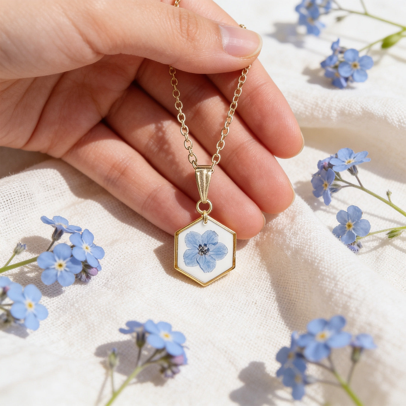 Hand holding a handmade botanical necklace for size reference, featuring a real forget-me-not flower on a white background. Surrounded by delicate blue forget-me-not flowers and soft linen in bright natural daylight with a light and airy summer atmosphere.
