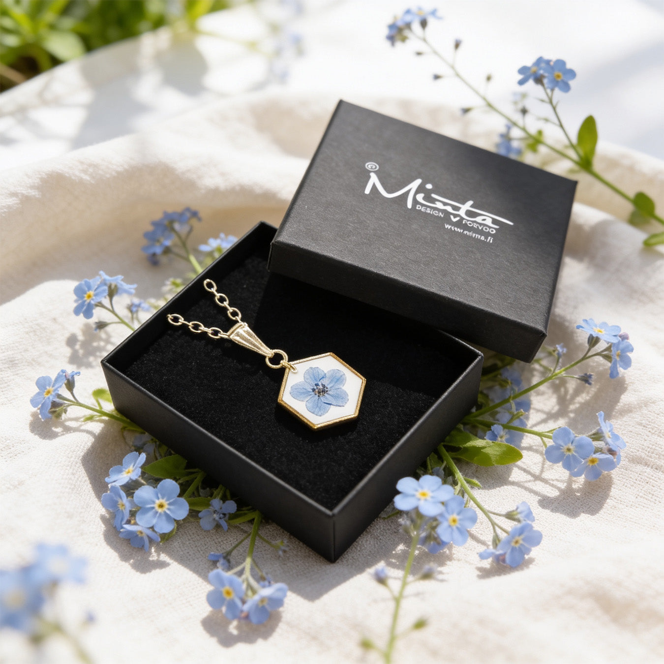 Handmade botanical necklace with a real forget-me-not flower displayed in a black jewelry box surrounded by delicate blue forget-me-not flowers and soft linen. Bright natural daylight and a light, airy summer atmosphere.