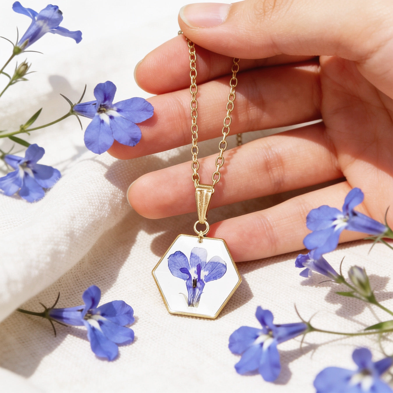 Hand holding a handmade botanical necklace by the chain for size reference, featuring a real blue lobelia flower on a white background. Surrounded by delicate blue lobelia flowers and light linen in bright natural daylight with a soft and airy summer atmosphere.