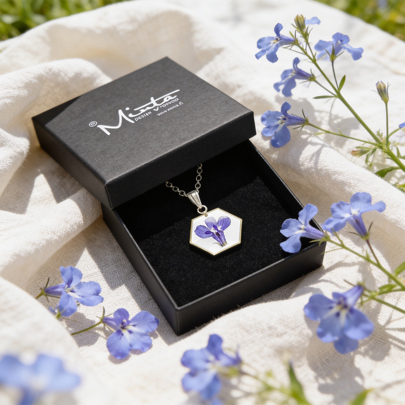 Handmade botanical necklace with a real blue lobelia flower displayed in a black jewelry box surrounded by delicate blue lobelia flowers and soft linen. Bright natural daylight and an airy summer atmosphere.