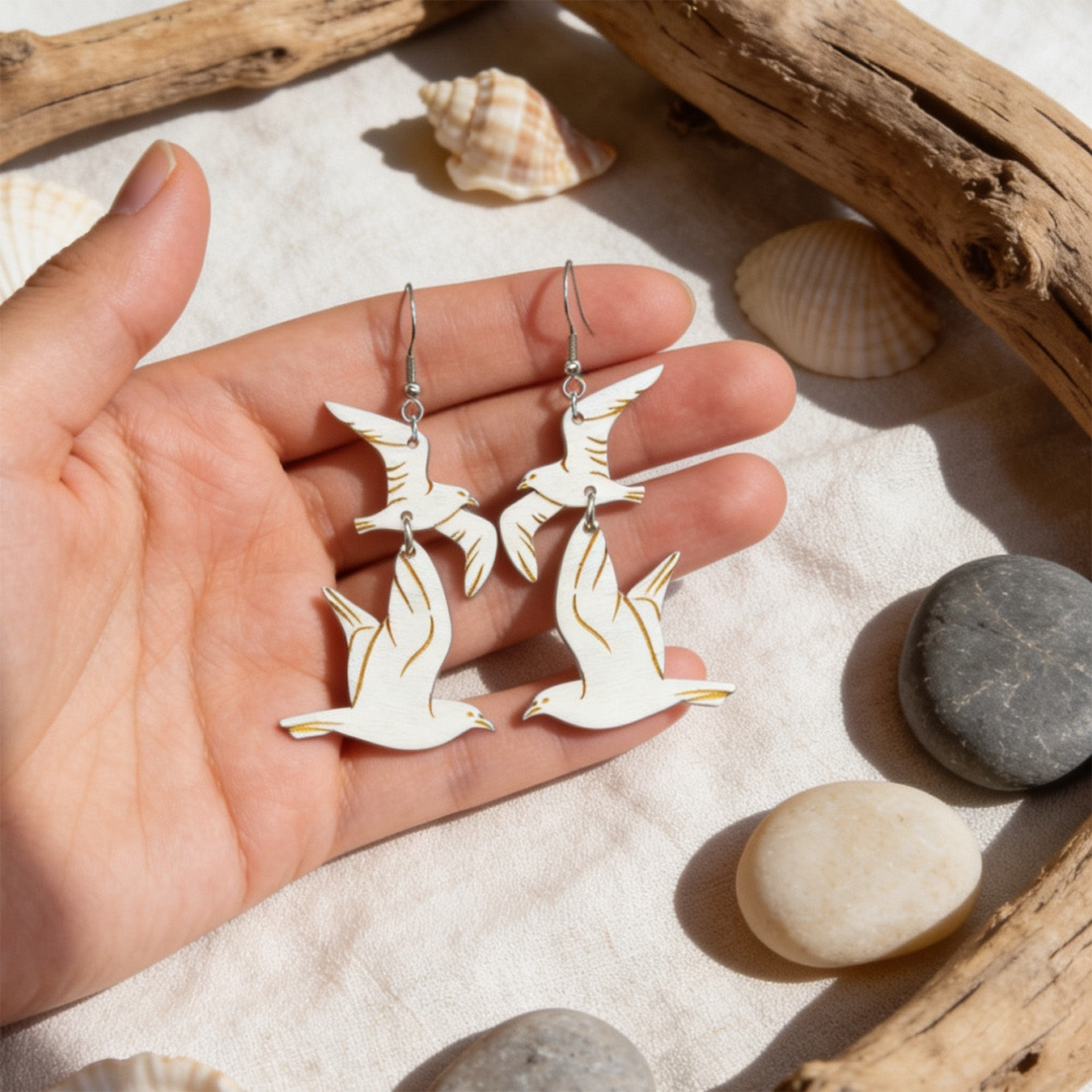 Hand holding handmade seagull earrings for size reference with driftwood, seashells and smooth beach stones. Lightweight Finnish birch plywood earrings photographed in soft natural sunlight with a fresh Nordic coastal atmosphere.