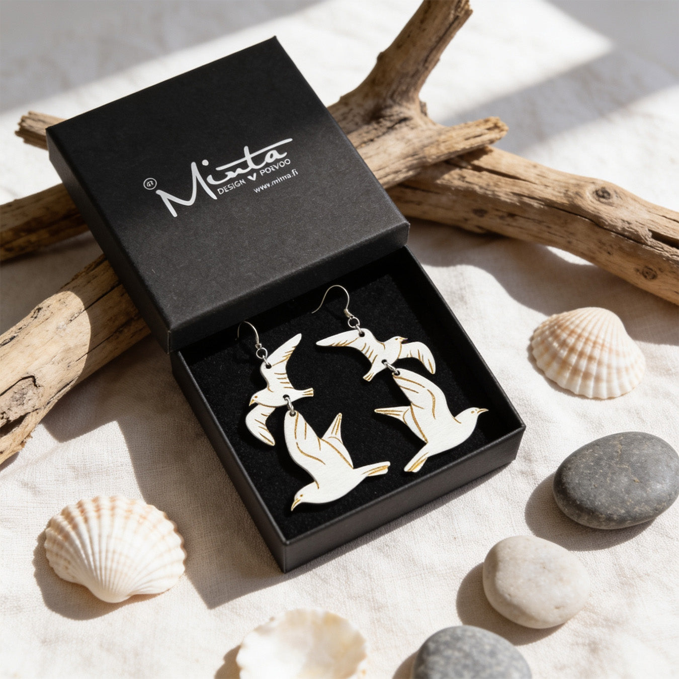 Handmade seagull earrings displayed in a black jewelry box surrounded by driftwood, seashells and smooth beach stones. Lightweight Finnish birch plywood earrings styled in soft natural daylight with a fresh Nordic coastal atmosphere.