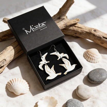 Handmade seagull earrings displayed in a black jewelry box surrounded by driftwood, seashells and smooth beach stones. Lightweight Finnish birch plywood earrings styled in soft natural daylight with a fresh Nordic coastal atmosphere.