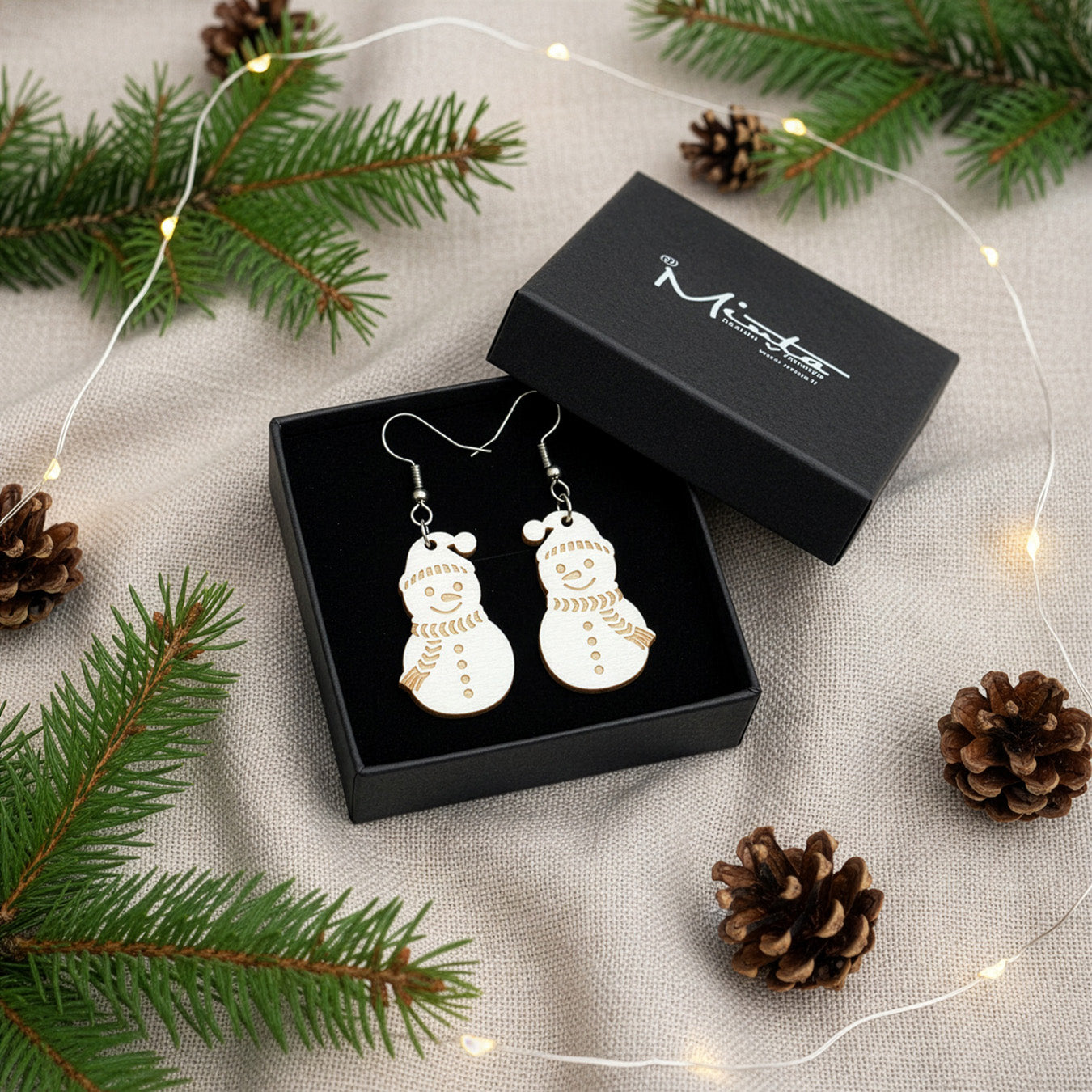 Snowman earrings presented in a gift box with pine branches, pinecones, and soft fairy lights, creating a festive Christmas atmosphere. Handmade in Finland by byMinta.